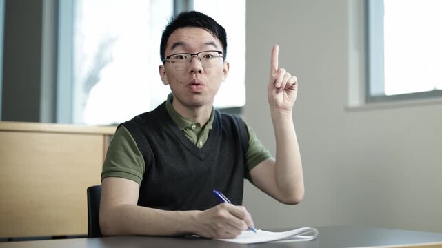 A young Asian male student wearing glasses raises his finger in a sudden moment of realization while writing an exam at a desk, ideal for student insight, problem solving, or academic breakthrough.