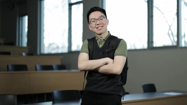 A young Asian male student wearing glasses and a backpack stands confidently with arms crossed and smiling in an empty lecture hall, ideal for student confidence, campus life, or academic success conc