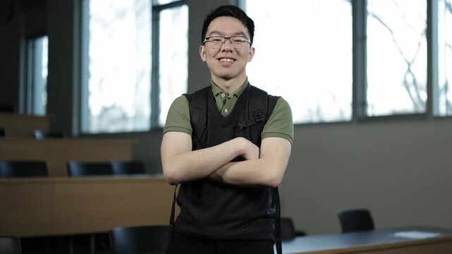 A young Asian male student wearing glasses and a backpack stands confidently with arms crossed and smiling in an empty lecture hall, ideal for student confidence, campus life, or academic success conc