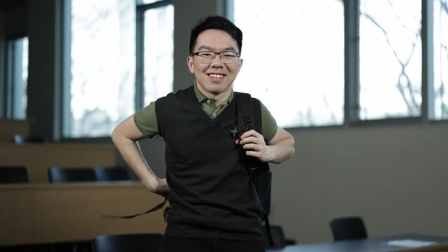 A young Asian male student wearing glasses smiles while putting on his backpack in an empty lecture hall, ideal for student life, back to school, or campus routine concepts.