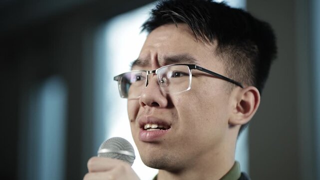 A close up of a young Asian male student wearing glasses showing a fearful, anxious expression while speaking into a handheld microphone, ideal for fear of public speaking, speech anxiety, or stage fr