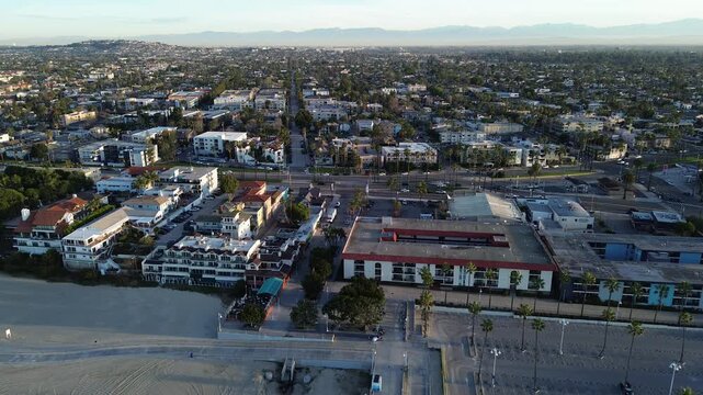 Backward flyover view Beachfront residential edge by Belmont Shore north of pier, East Ocean Blvd connects dense residential blocks to coast, lifeguard tower positioned along sand, Long Beach, CA