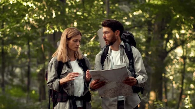 A man with light skin and a woman with light skin are looking at a map while hiking in a green forest. They are dressed in outdoor gear and backpacks. Sunlight filters through the trees.