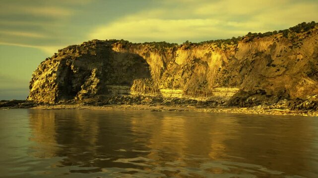 sunlit bluff and calm sea with shimmering reflections, wet sand and scattered stones add foreground texture, soft sky enhances peaceful evening mood