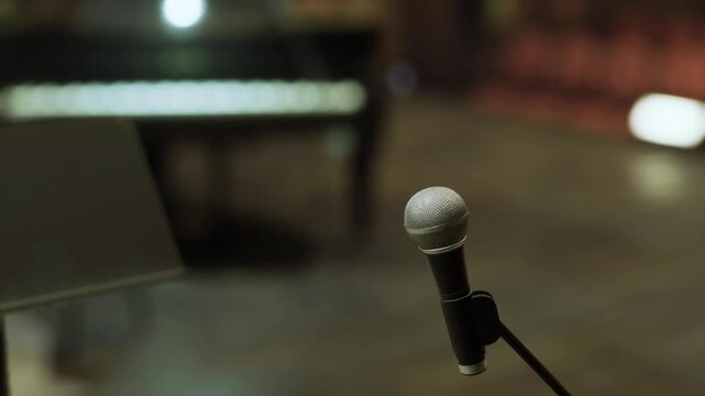 Ready microphone at empty stage for spoken word performance, solitary spotlight, blurred piano suggesting live accompaniment, quiet anticipation, audience seats
