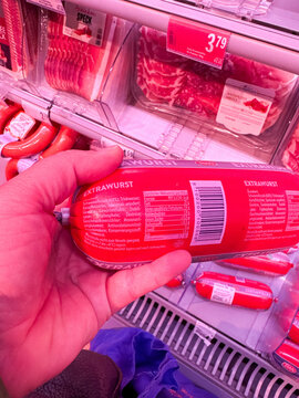 Villach, Austria - January 29, 2026: Hand holding a red package of extrawurst sausage in a refrigerated display case with various meat products visible in the background
