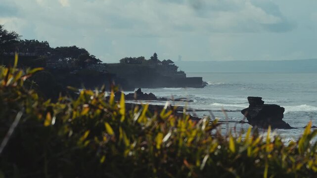 Rock Formation Framed By Surf,Towering Sea Stack With Pounding Waves,Mist Veil And Fierce Spray,Distant Temple Pagoda Tanah Lot On Cliff, Moody Light, Geology And Erosion Focus For Travel Documentary.