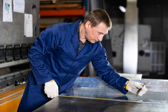 Serious middle-aged technician in blue jacket and protective gloves measuring steel sheet with tape measure in industrial setting of metalworking workshop, focused on ensuring accuracy of measurements