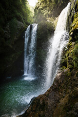 Obraz premium Waterfall Flowing Over Mossy Rocks in a Forest with Sunlight Filtering Through the Trees
