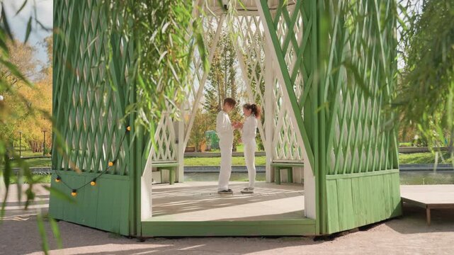 White couple exchanging vows in gazebo, green lattice arbor framed by willow branches and autumn foliage, bride in white dress and groom in light suit sharing tender embrace and kiss, ring exchange
