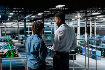 Server hub teamworking IT experts visualizing large language model architecture using laptop on elevated industrial platform. Data center system administrators collaborating on tasks.