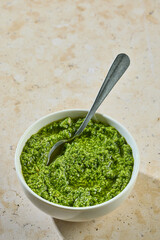 Fresh homemade pesto in white bowl with spoon on sunny kitchen table