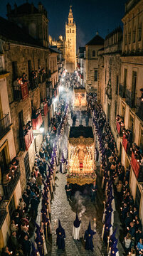 Procesi&oacute;n nocturna de Semana Santa en Espa&ntilde;a con nazarenos de t&uacute;nicas moradas y velas encendidas, acompa&ntilde;ando un paso iluminado por cirios en calle hist&oacute;rica empedrada.