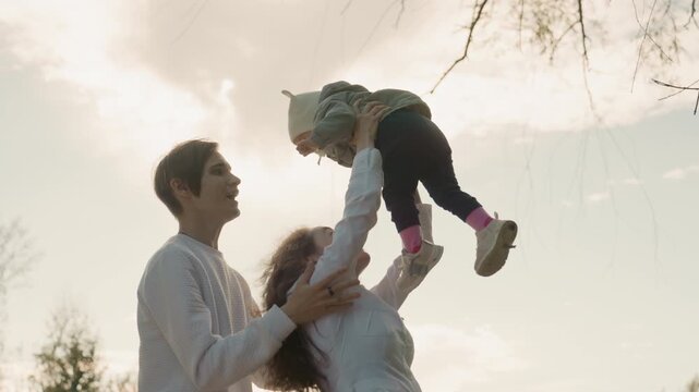 Parents toss baby into sky under sunset, exuberant family and marriage celebration with both parents laughing, baby wearing hat and boots, golden backlight, playful high toss and spin