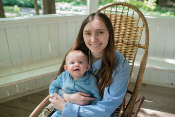 Fototapeta premium Happy young woman holding baby boy while sitting in wicker rocking chair on white porch