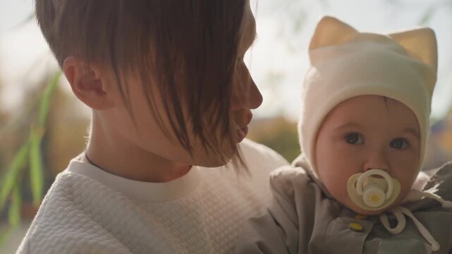 Shoreline closeup protective parent with baby, pacifier and cozy hat visible, contemplative gaze over calm water, soft backlight and autumn palette convey quiet emotional bond and nurture.