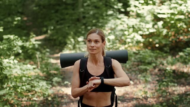 A woman with light skin walks on a forest trail while checking her watch. She carries a yoga mat on her back and enjoys the outdoors. Sunlight filters through the trees along the path.