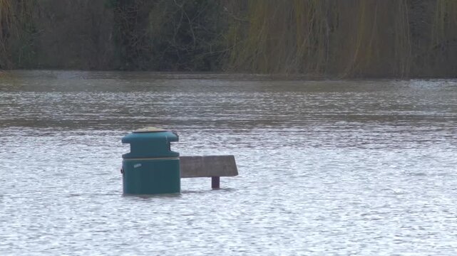 park bench and rubbish bin aka trash can in flooded city park, poking through the water, trees in background
