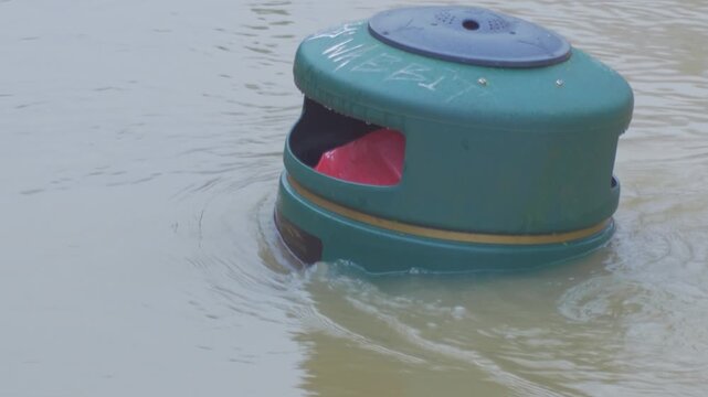 flood water nearly covering rubbish bin aka trash can, top of bin visible, surrounded by fast flowing muddy water, flooded city park