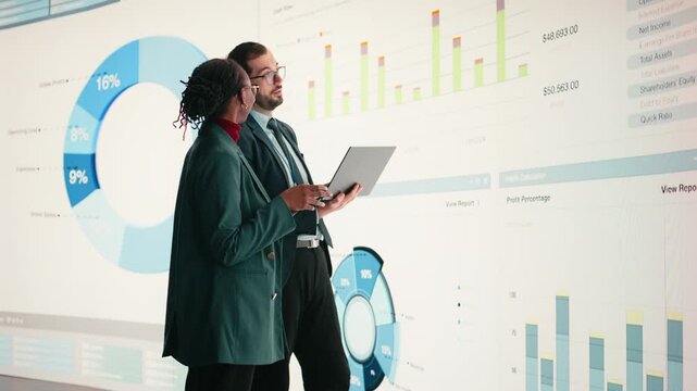 Corporate managers evaluate fiscal performance metrics on digital wall, collaborating. Diverse executive team discusses quarterly earnings report and shareholder equity charts during boardroom meeting