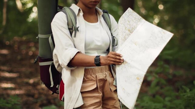 A woman with dark skin studies a map in a dense forest. She has a backpack and a camping mat. Sunlight filters through the trees, highlighting her focused expression as she plans her route.