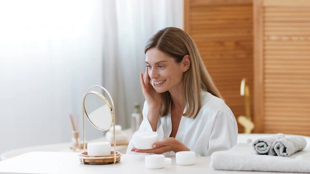 A woman with long hair sits at a vanity table in a modern bathroom. She is applying skincare cream while looking in the round mirror. Towels and small jars are nearby.