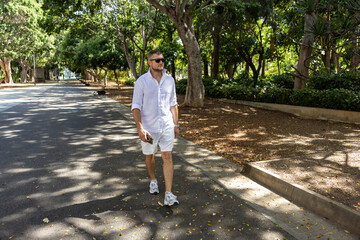 Casual Man Walking In Green Urban Park On Sunny Summer Day With Smartphone
