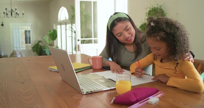Diverse female mom leaning, pointing at paper and laptop, guiding child girl writing for schoolwork