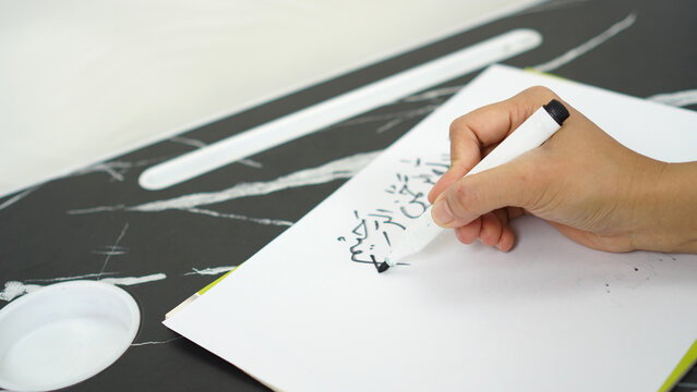 a woman is writing the Arabic sentence Basmalah or Bismillahirrahmanirrahim which has the meaning of saying the name of Allah, the Most Gracious and Most Merciful, using a black marker on white paper