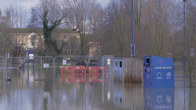 flooded riverside car park, recycling bins, fence, buildings in background, reflections, lamp posts