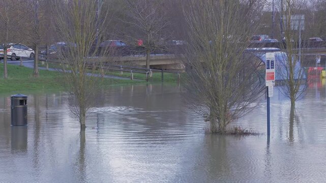 flooded riverside car park, road bridge with traffic in background, submerged trees, bin and signposts