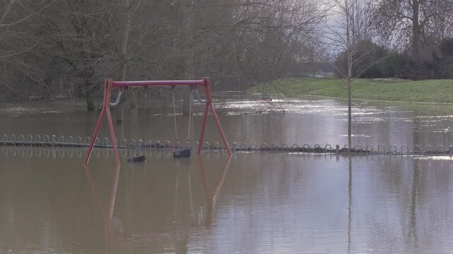 Flooded playground, chidren's play area, swings submerged in flood water, embankment, trees in background