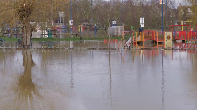 Flooded park, children's play area, swings and climbing frame submerged in flood water, cars in distance. Reflection of large tree and lamp posts in foreground