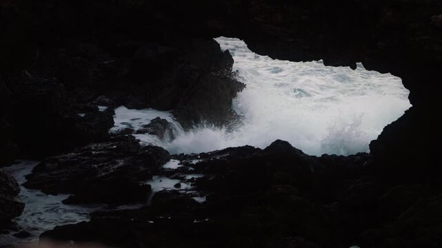 Powerful Mediterranean waves crashing against dark rocky coastline in Ibiza filmed from natural rock opening creating dramatic coastal scene.