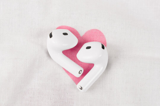 Two white wireless earbuds arranged into a heart shape on white textured fabric with pink heart beneath, romantic tech concept for music lovers with minimal clean layout and copy space