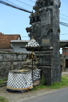 Stone Statue of Balinese Deity Beside Traditional Candi Bentar Gate on Road with Ritual Umbrella and Sacred Cloth