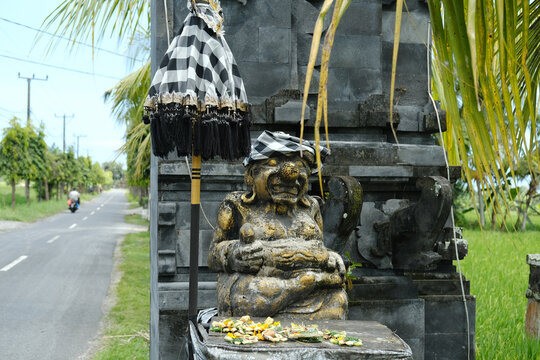 Stone Statue of Balinese Deity Beside Traditional Candi Bentar Gate on Road with Ritual Umbrella and Sacred Cloth