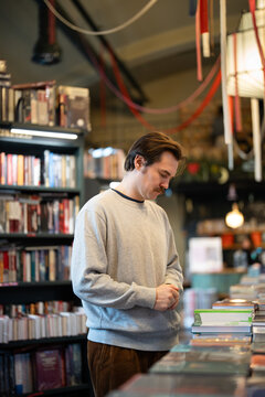 Focused interested man picking books in modern bookshop on table display, looking at stacks of publications, reading titles and covers. Pondering male booklover thinking on book to buy at book fair.
