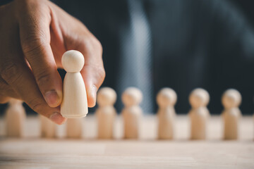 A hand holding a small wooden figure in front of a row of similar figures. The background is blurred.