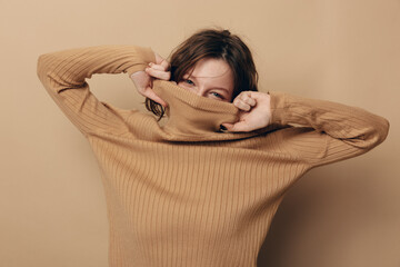 Young woman in beige knitted sweater pulls collar over face, smiling with playful eyes. Neutral...