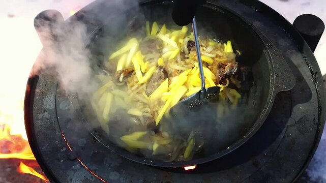 Gloved hand stirring food in pan over open flame. Survival skills, camping activity and rustic outdoor preparation. Cooking of pilaf with beef meat and carrot.