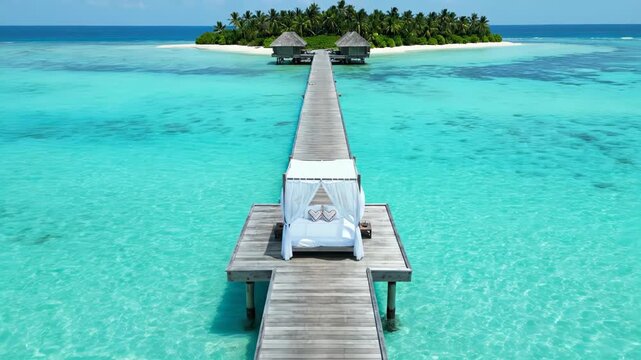 A romantic canopy bed sits on a pier over clear turquoise water. The pier leads to a beautiful tropical island with palm trees.