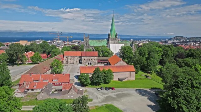 Aerial view of Nidaros Cathedral in Trondheim, Norway, Norwegian rooftops and leafy grounds framing the historic complex . Drone view glides closer, tightening on the copper-green spire above the city