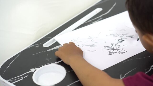 Asian little boy is learning to write and scribble on white paper using a black marker to learn to write letters before school