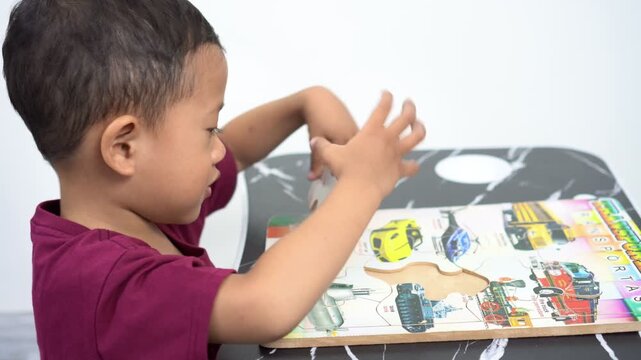 A small Asian boy is playing, focusing, concentrating on putting together a puzzle with pictures of land transportation on a black table.
