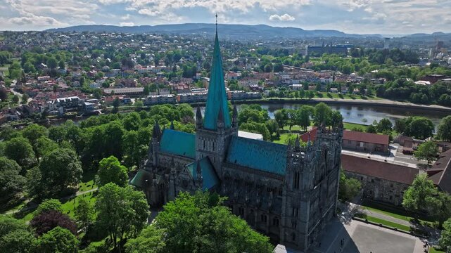 Aerial view of Nidaros Cathedral in Trondheim, Norway, Norwegian treetops surrounding the nave and transept . Drone view eases past the spire, opening the frame to the river bend and city blocks.