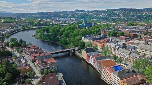 Aerial view of Trondheim and the Nidelva River, Norway. Drone glides over riverside streets, bridges, and historic buildings with green parks and clear summer daylight.