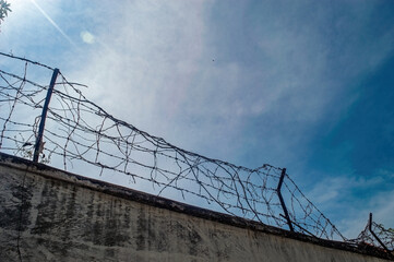 A rugged concrete wall is topped with tangled barbed wire under a bright, cloudy sky, conveying themes of security, isolation, or urban decay