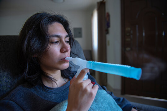 Mujer joven hispana Mexicana en tratamiento de nebulizacion en casa por problemas respiratorios de bronquitis y asma. Vista lateral