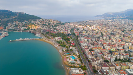 Fototapeta premium Alanya, Turkey. Municipal beach with surf and umbrellas, seafront hotels, marina and coastal road with cars in Alanya summer season.. Aerial View
